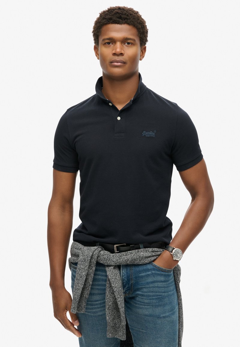 Marineblauw poloshirt met een geknoopte kraag, voorzien van een subtiel logo. Draagt bij lichte blauwe jeans en een grijze trui geknoopt om de taille.