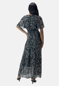 Schwarzes Blumenkleid im Maxistil aus halbtransparentem Material, mit elastischem Bund, flatternden Ärmeln und weißem Blumenmuster. Die Rückansicht zeigt die fließende Silhouette.