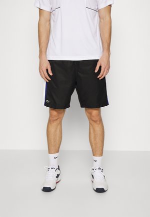 Mężczyzna w czarnych sportowych szortach, białych skarpetkach Nike, białych butach sportowych i białej koszulce z krótkim rękawem na jednolitym tle.