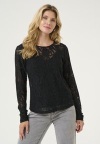 Top lungo in pizzo nero con maniche, caratterizzato da un motivo floreale, scollatura rotonda e polsini aderenti, abbinato a jeans grigi. Tessuto strutturato e semitrasparente.