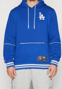 Felpa blu con lacci bianchi, caratterizzata da un logo frontale e inserti a righe ai polsini e all'orlo. Il materiale appare morbido e textured.
