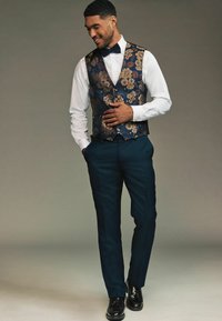 Anzug mit dunkelblauen Hosen und einer gemusterten Weste aus marineblauem und goldenen Blumenstoff, kombiniert mit einem weißen Hemd und einer Fliege.