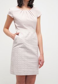 Robe à manches courtes en rose pâle avec un corsage structuré, des plis au niveau de l'encolure et une jupe à motifs présentant des formes géométriques.