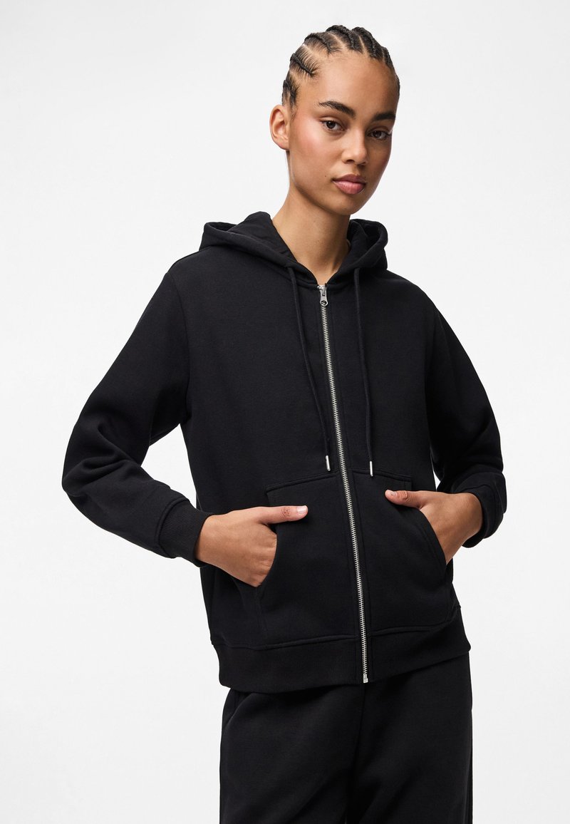 Kvinna med flätat hår som bär en svart zip-up hoodie med händerna i framfickorna, stående mot en enfärgad vit bakgrund.