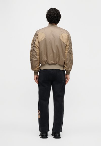 Μπουφάν bomber από μπεζ νάιλον με ριμπ μανσέτες και ζώνη στη μέση· διαθέτει υφασμένους τονισμούς στους ώμους και χαλαρή γραμμή. Συνδυάζεται με μαύρο παντελόνι.