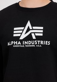 Felpa nera con logo grafico bianco a forma di "A" e tre linee orizzontali. Il testo recita "ALPHA INDUSTRIES, KNOXVILLE, TENNESSEE, U.S.A."
