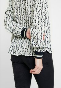Blouse blanche avec un motif tourbillonnant noir et vert, dotée de poignets ribbed noirs avec des accents verts, en tissu à texture douce, et avec un ourlet froncé.