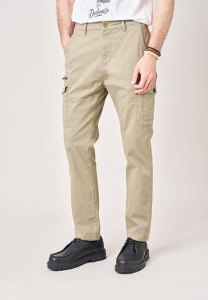 Man die beige cargobroek met zijzakken en zwarte veterschoenen draagt, staat met handen in de zakken tegen een effen achtergrond.