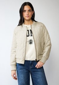 Veste bomber matelassée beige avec fermeture éclair et poignets côtelés. Portée par-dessus un T-shirt graphique de couleur claire et associée à un jean bleu.