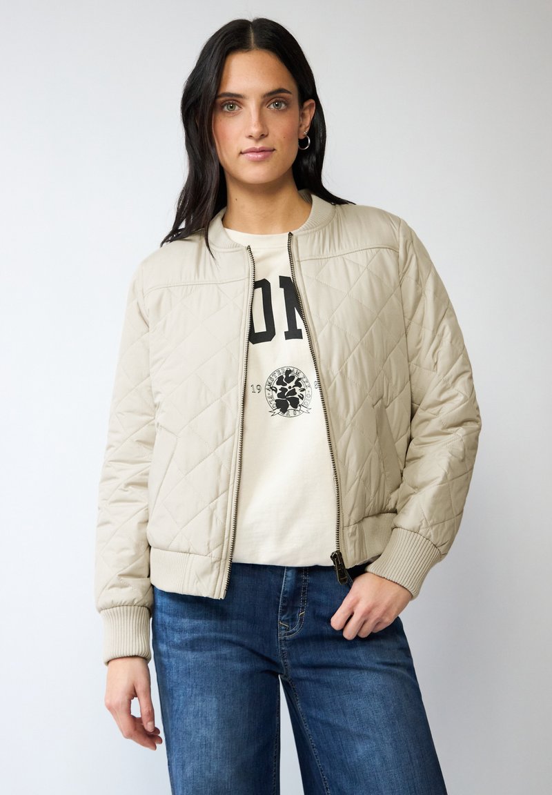 Veste bomber matelassée beige avec fermeture éclair et poignets côtelés. Portée par-dessus un T-shirt graphique de couleur claire et associée à un jean bleu.