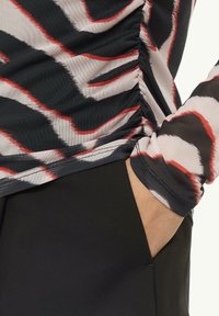 Langärmliges Oberteil in einem abstrakten Muster aus Schwarz, Weiß und Rot mit gerafftem Seitendetail; kombiniert mit schwarzen Hosen, wobei eine Hand in der Tasche zu sehen ist.