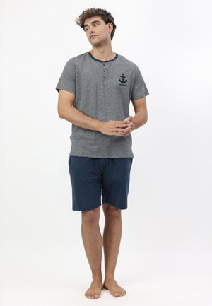 Joven de pie descalzo, vistiendo una camiseta Henley gris de manga corta con logo de ancla y pantalones cortos azul marino, sobre un fondo blanco.