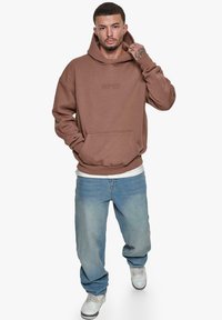 Brun hætte sweatshirt med en frontlomme, der har diskret mærkeschrift. Sammen med lyseblå baggy jeans og hvide sneakers.