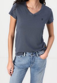 Šedé v-neck tričko vyrobené z bavlny s krátkými rukávy, vystavené s světle modrými džíny. Jemné logo na hrudi.