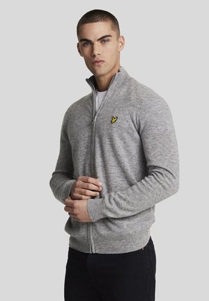 Pull gris zippé en tissu tricoté doux. Il présente un col et des poignets côtelés, avec un petit logo jaune sur la poitrine.