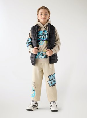 Giovane in piedi che indossa un gilet imbottito nero sopra una felpa beige con grafica e pantaloni della tuta abbinati con scarpe da ginnastica bianche.