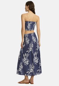 Modelo vistiendo un conjunto de dos piezas con estampado floral, que incluye un top corto sin tirantes y una falda larga, visto desde la espalda. Sandalias de color arena visibles.