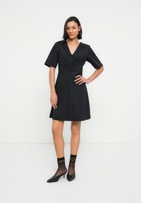 Μαύρο φόρεμα με κοντά μανίκια, V-λαιμόκοψη και ρυτιδωμένη λεπτομέρεια στο μπροστινό μέρος. Φορεμένο με διάφανα μαύρα καλτσάκια και μαύρες γόβες με μυτερή μύτη.