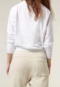 Persona con capelli lunghi e ondulati, vestita con una camicia bianca a maniche lunghe e pantaloni felpati beige, vista di spalle su uno sfondo semplice.