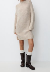 Beżowa dzianinowa sukienka swetrowa o luźnym kroju, z bocznymi rozcięciami i długimi rękawami. Połączona z brązowymi skórzanymi kozakami sięgającymi kolan oraz białymi skarpetkami za kostkę.