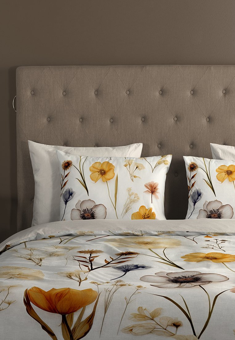 Bett mit beigem gepolstertem Kopfteil, weißer Bettwäsche mit gelben, braunen und blauen Blumenmustern auf Decke und Kissen, vor einer braunen Wand.