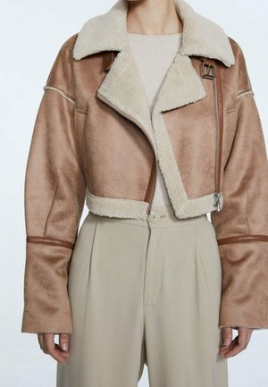 Giacca corta marrone in shearling con ampio colletto e cerniera, indossata sopra un maglione beige e pantaloni beige a vita alta.