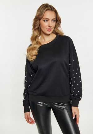 Frau mit langem, welligem Haar, die einen schwarzen Pullover mit Perlendetails an den Ärmeln und schwarze Lederhosen trägt und zur Seite schaut.