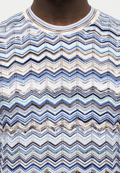 Κοντινό πλάνο ενός ατόμου που φοράει πλεκτή μπλούζα με ζιγκ-ζαγκ μοτίβα σε μπλε, λευκό, καφέ και τιρκουάζ χρώμα και στρογγυλή λαιμόκοψη.