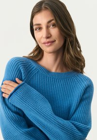 Blå strikket sweater med ribbet tekstur; har vid halsudskæring og lange ærmer. Modellen poserer med hænderne krydset over torsoen.