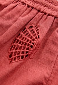 Rødt tekstureret stof med en rynket talje har et detaljeret lattice broderiudskæring, der tilføjer visuel interesse og et unikt designelement.