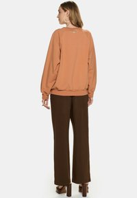 Brązowa bluza z długim rękawem, z ribbowanym wykończeniem u dołu i detalem logo na plecach, połączona z ciemno-brązowymi spodniami w szerokim kroju i sandałami na słupku.
