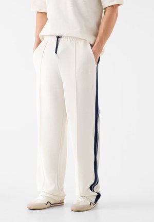 Person trägt weiße, strukturierte Hose mit dunkelblauen Seitenstreifen, einem Kordelzug in der Taille und weißen Sneakern mit beigen Akzenten.