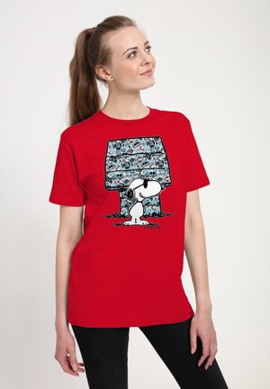 Femme portant un t-shirt rouge avec Snoopy portant des lunettes de soleil, debout devant une niche bleue couverte de texte style graffiti.