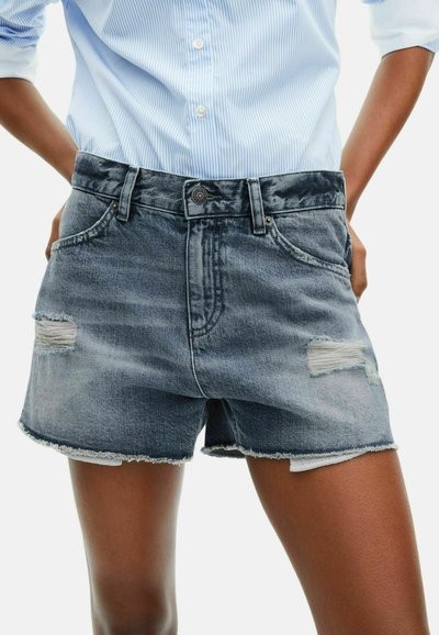 Gros plan d'une personne portant une chemise rayée bleu clair et un short en denim déchiré avec des bords effilochés, les mains reposant sur les hanches.