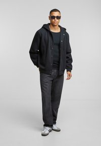 Zwarte hoodie met drukknoopjes, gecombineerd met een zwarte tanktop en donkere spijkerbroek. Schoeisel heeft een grijs-wit ontwerp met strepen.