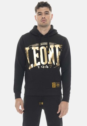 Uomo che indossa una felpa nera con cappuccio con scritta dorata "LEONE 1947" e pantaloni neri abbinati, in piedi su sfondo bianco, rivolto verso la fotocamera.