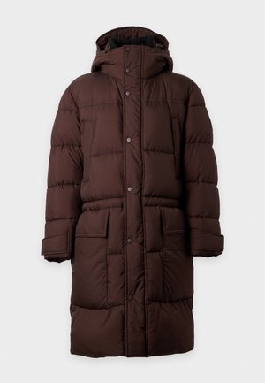 Un homme porte un long manteau matelassé marron foncé sur un haut marron, un jean bleu clair à jambes larges et des chaussures noires, sur un fond blanc uni.