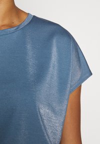 Modrý top s krátkým rukávem a jemnou, lesklou texturou. Má kulatý výstřih a volný střih s hladkým materiálem, který odráží světlo.