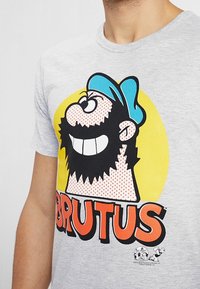 Szara bawełniana koszulka z grafiką przypominającą postać z kreskówki w niebieskiej czapce, z dużym uśmiechem, czarną brodą oraz napisem "BRUTUS" w grubym pomarańczowym kolorze.