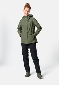 Giacca impermeabile verde con cappuccio, tasche laterali e zip frontale. Abbinata a pantaloni neri e scarponi da trekking verdi. Texture liscia.