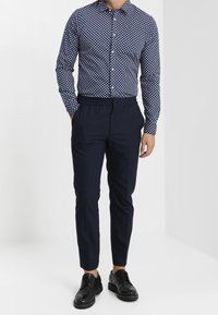 Man in marineblauwe broek, zwarte nette schoenen en een marineblauw overhemd met witte stippen, staand tegen een witte achtergrond.