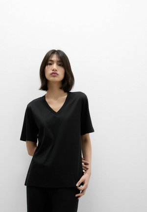 Mladá žena s tmavými vlasy po ramena stojí před prostou bílou zdí, oblečená v černém tričku s výstřihem do V a černých kalhotách.