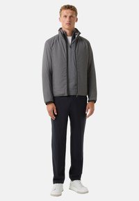 Grå zip-up jakke med en tekstureret overflade, parret med en grå skjorte og mørke bukser. Modellen har hvide sneakers på. Enkel, moderne design.