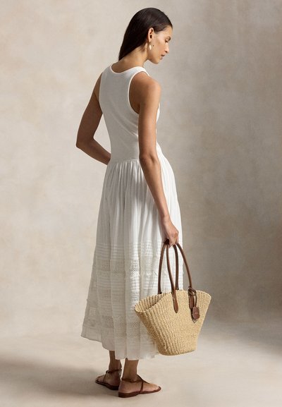 Naine, kes kannab pitsiliste detailidega valget varrukateta kleiti, hoiab käes kootud õllekõrte käekotti ning jalas on tal pruunid lamedad sandaalid, seisab külje pealt.