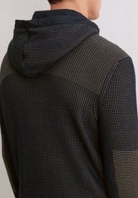 Mann in schwarzer und beigefarbener strukturierter Strickjacke, von hinten vor einem schlichten hellen Wandhintergrund dargestellt.