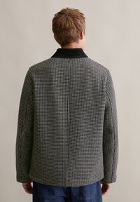 Grijs houndstooth-patroonjas met een zwarte fluwelen kraag, rechte snit en lange mouwen, gefotografeerd vanuit de rug.