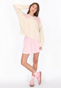 Crème gebreide cardigan met roze bloemenapplicaties op de schouders, gecombineerd met een lichtroze geplisseerde rok, roze sneakers en enkelkousen.