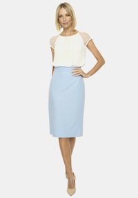 Biała bluzka z przezroczystymi rękawami w kropki, zestawiona z jasnoniebieską spódnicą ołówkową. Modelka stoi w neutralnych szpilkach, z rękami na biodrach.