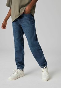 Jeans de mezclilla azul oscuro con un ajuste relajado, que presentan un diseño clásico de cinco bolsillos y un desvanecimiento sutil, combinados con zapatillas blancas.