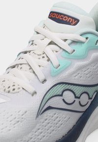 Λευκό παπούτσι τρεξίματος Saucony με λευκά κορδόνια, τιρκουάζ και ναυτικό μπλε λεπτομέρειες, και αεριζόμενο ύφασμα από πλέγμα.